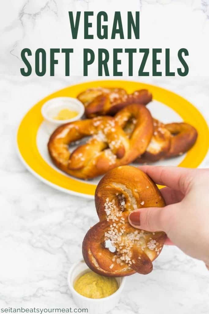Hand dipping a salted soft pretzel in a cup of mustard with text "Vegan Soft Pretzels"