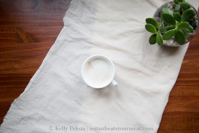 Overhead photo of mug of coffee with cashew milk on white tablecloth on wooden table