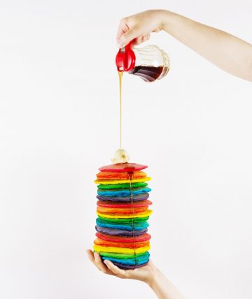 Hand holding tall stack of rainbow pancakes with other hand pouring syrup on them