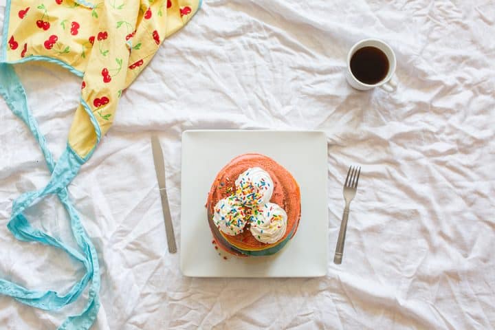 Overhead image of rainbow pancakes on a white plate on a white tablecloth with coffee an an apron