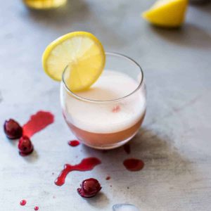 Close up photo of whiskey sour on silver counter surrounded by cherries and lemons