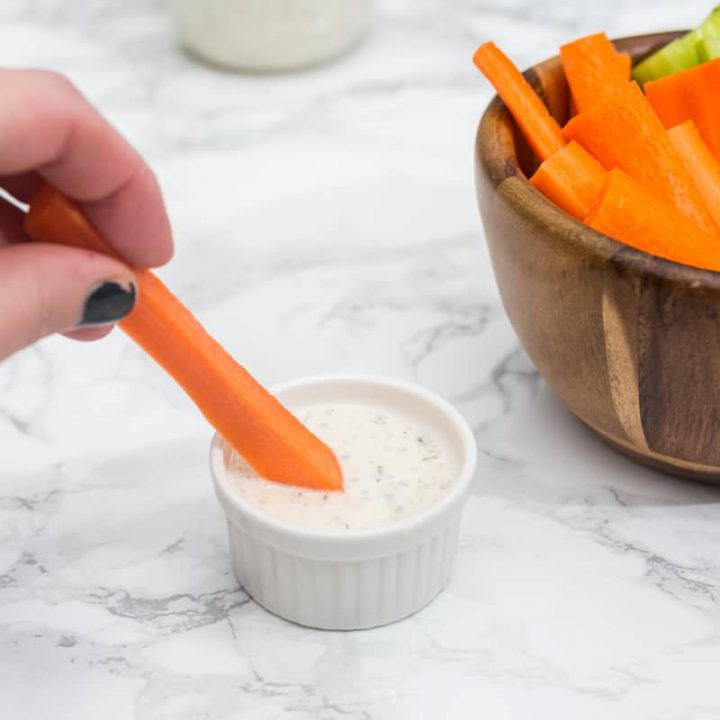 Hand dipping carrot stick in small cup of ranch dressing