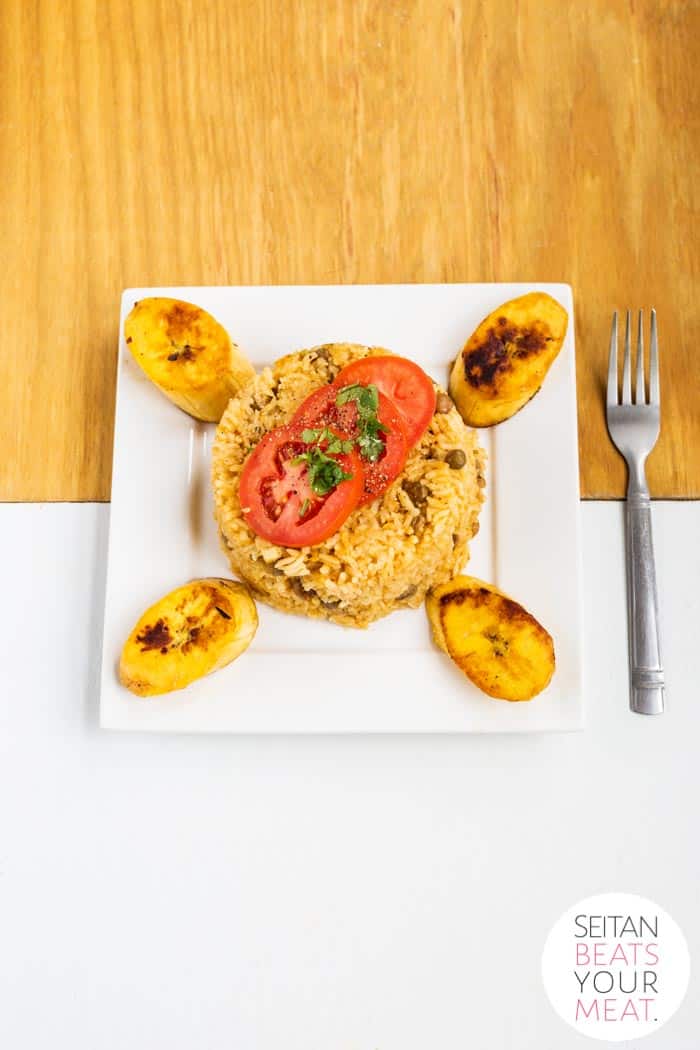 Overhead image of arroz con gandules with tomatoes and plantains on a white plate on wood background
