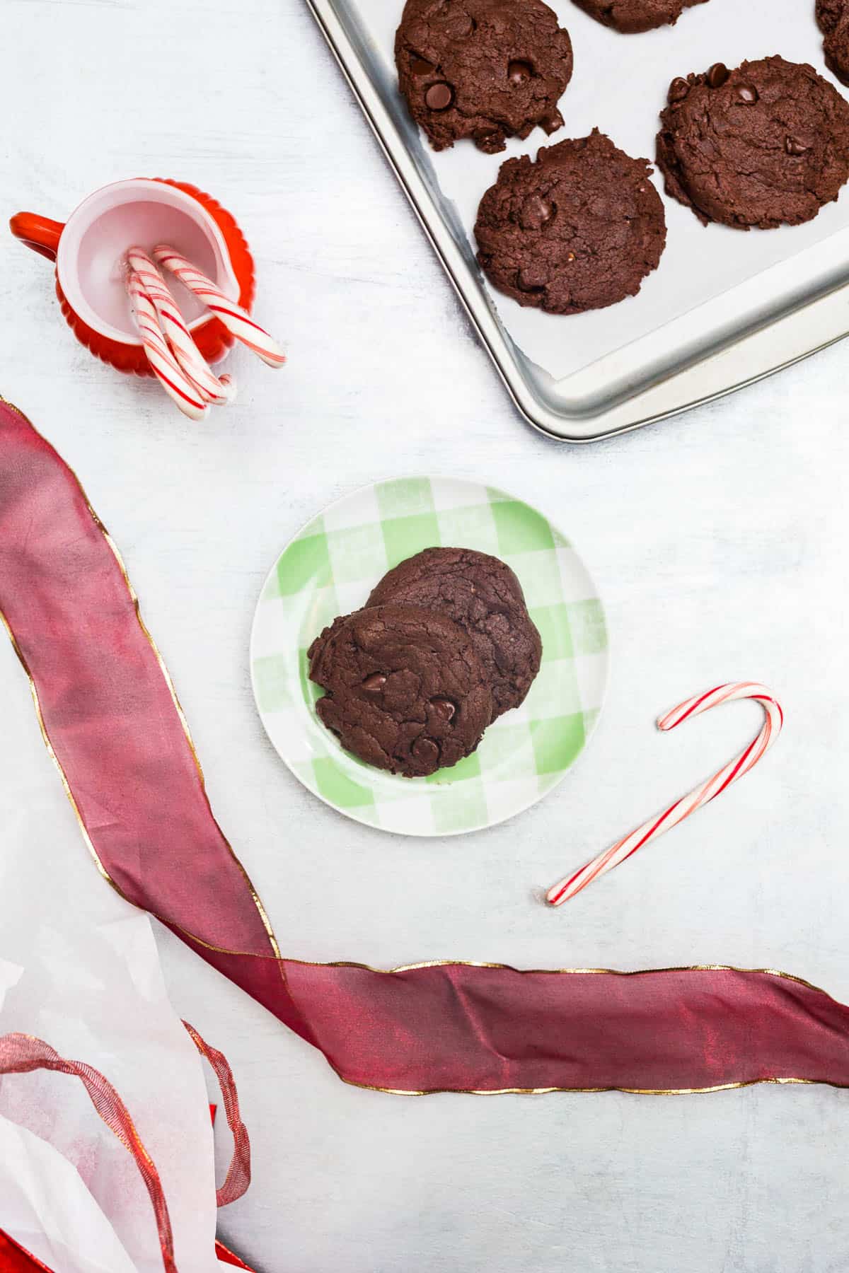 Chocolate cookies on green plate surrounded by red ribbons, gift bags, and candy canes on silver background