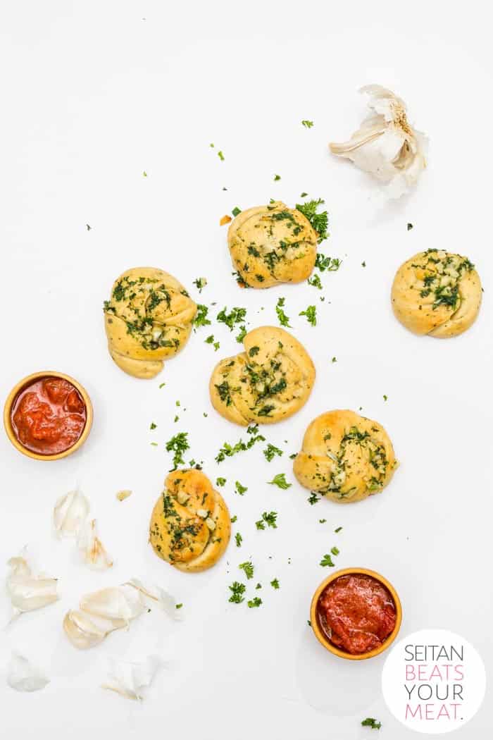 Overhead image of garlic knots with heads of garlic, marinara in cups, and chopped parsley