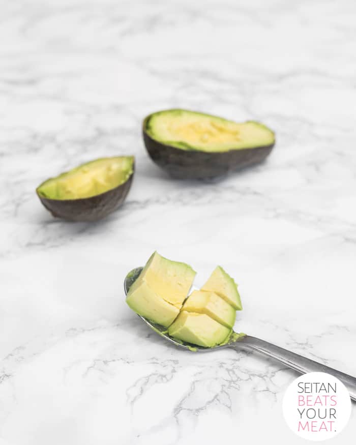 Cut avocado on spoon with avocado halves in background on marble surface