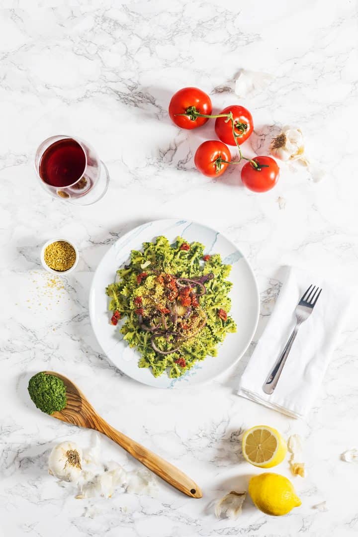 Kale pesto pasta on plate on marble counter surrounded by tomatoes, garlic, and lemons