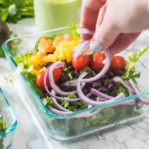 Hand adding red onion to salad in glass container on marble counter