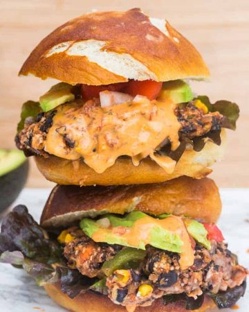 Stack of two black bean burgers on pretzel buns with avocado in background and chipotle mayo dripping down