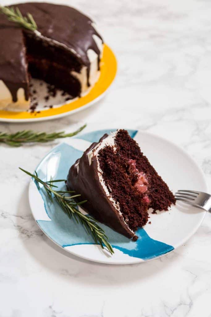 Slice of chocolate cake on plate with sprig of rosemary and rest of cake in background