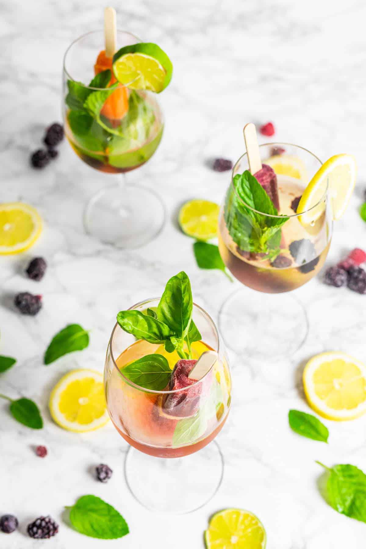 Three glasses of wine with a popsicle, lemon and lime slices, and fresh herbs