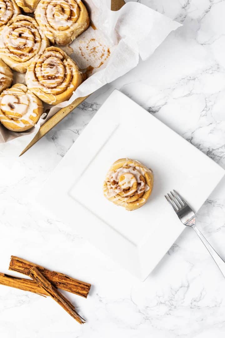 Cinnamon roll on plate with fork and tray of cinnamon rolls off to side