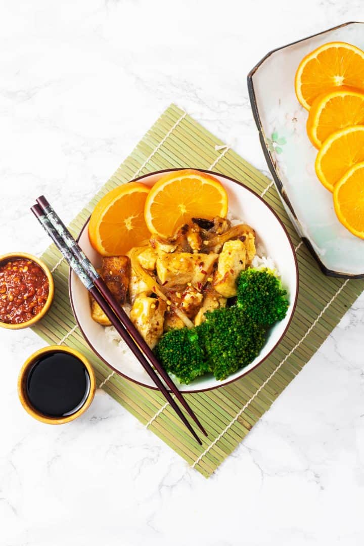 Bowls of orange tofu with broccoli on table setting with chopsticks