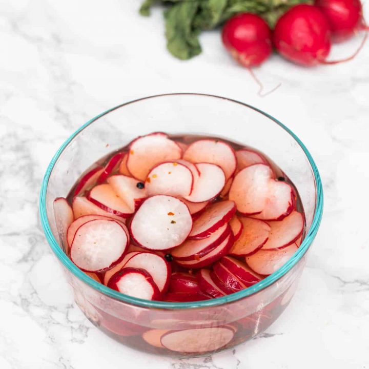 Glass bowl of radish slices in brine with pepper flakes with bunch of radishes on side