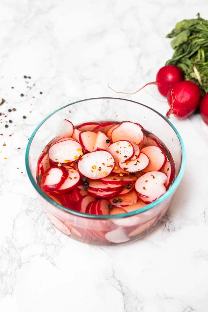 Glass bowl of radish slices in brine with pepper flakes with bunch of radishes on side