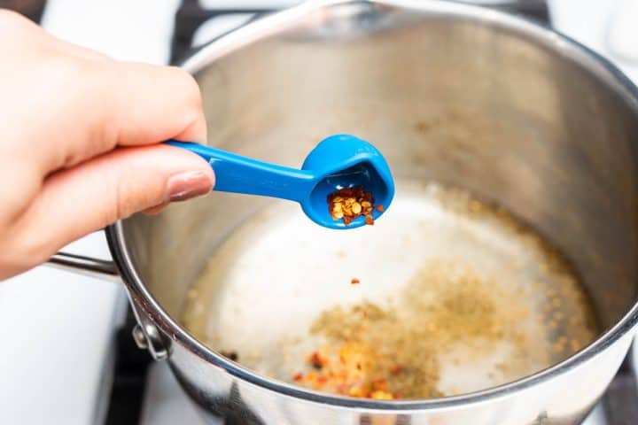 Hand pouring spoonful of chili flakes into pot on stove