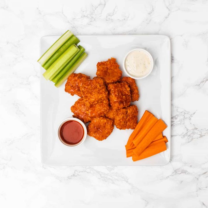 Overhead view of plate of buffalo tofu wings on plate with celery sticks, carrot sticks, ranch dressing, and buffalo sauce