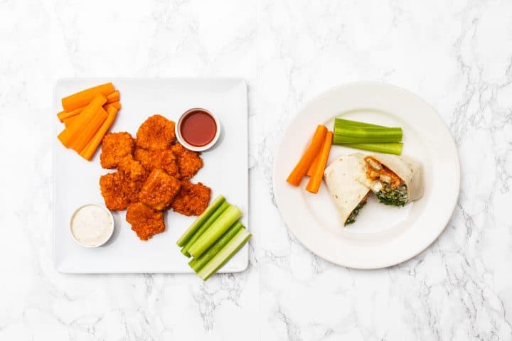 Overhead view of two plates beside each other, one with buffalo tofu wings, carrot and celery sticks, and sauces, and the other with a buffalo tofu wrap and carrot and celery sticks