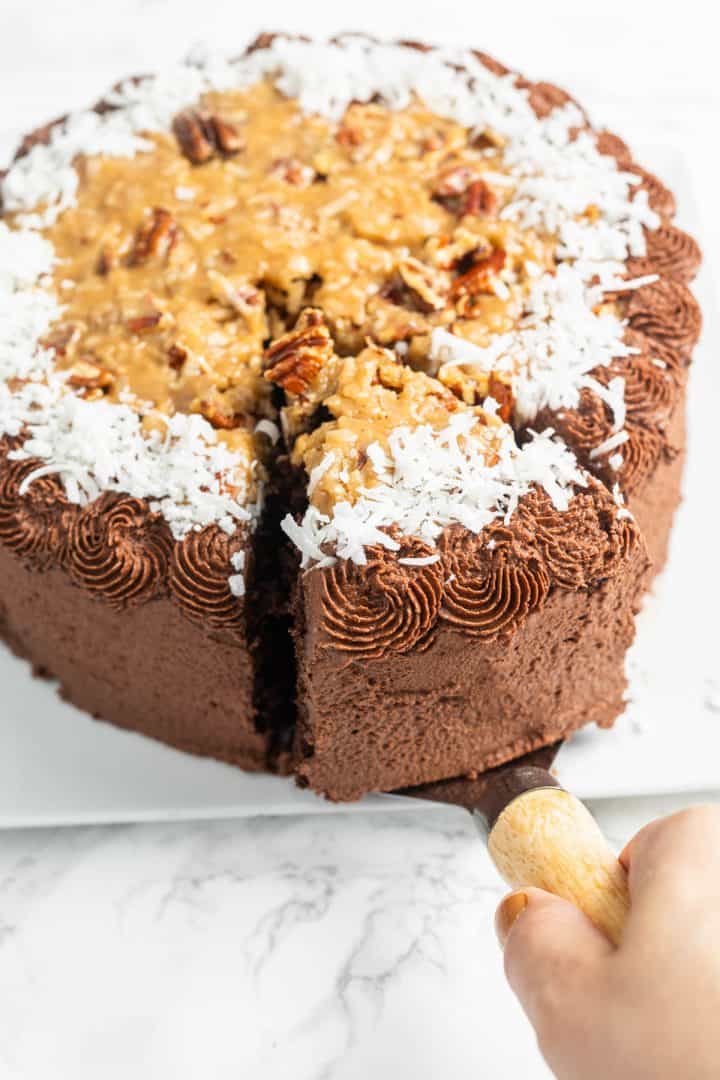 Hand lifting slice of chocolate cake with pecan coconut caramel and chocolate frosting with serving utensil