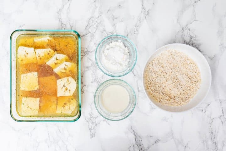 Overhead view of tofu in marinade in glass dish, small bowls of cornstarch and milk, and breadcrumbs in medium bowl