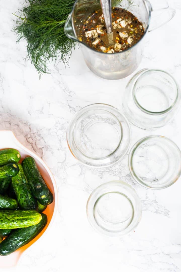 Four empty glass jars with pickle brine, pickling cucumbers, and fresh dill off to side