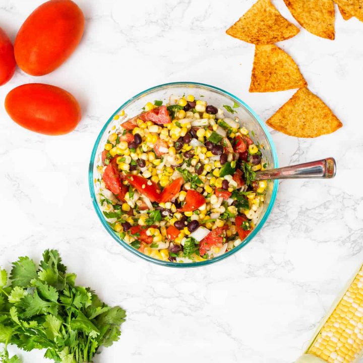 Bowl of corn and black bean salsa in glass bowl surrounded by ingredients
