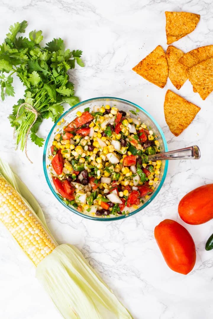 Bowl of corn and black bean salsa in glass bowl surrounded by ingredients