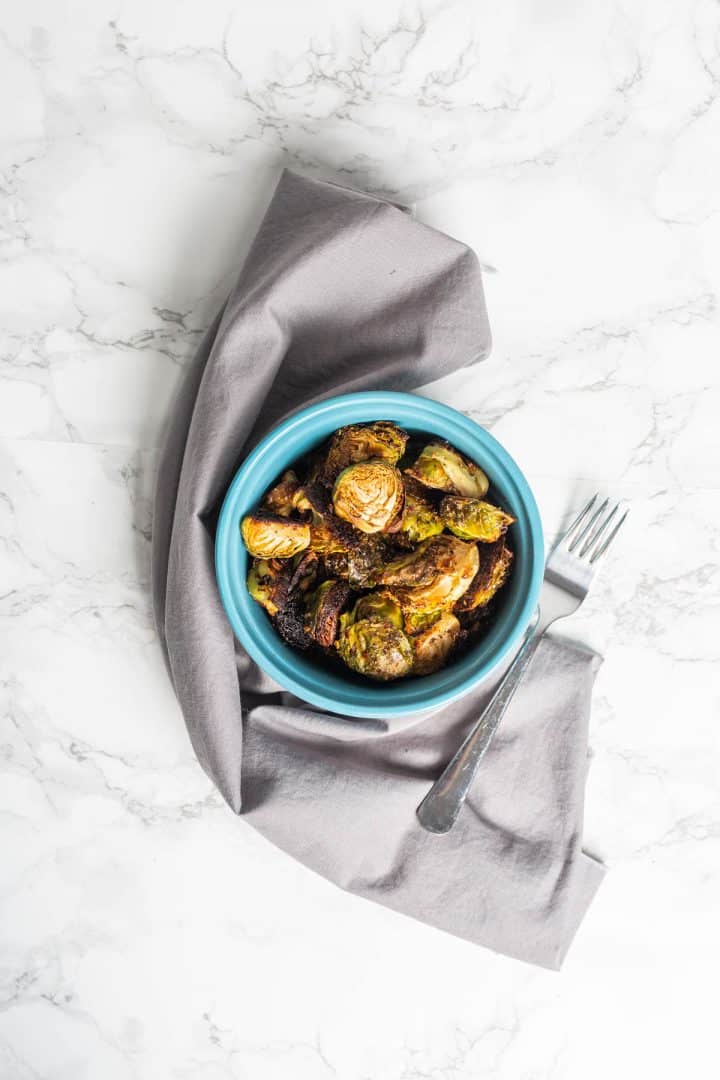 Bowl of roasted Brussels sprouts wrapped in gray cloth napkin with fork on marble counter