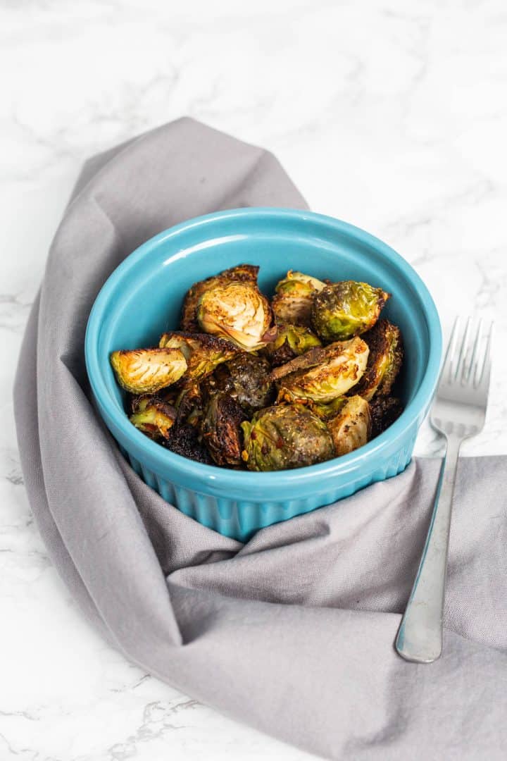 Bowl of roasted Brussels sprouts wrapped in gray cloth napkin with fork on marble counter