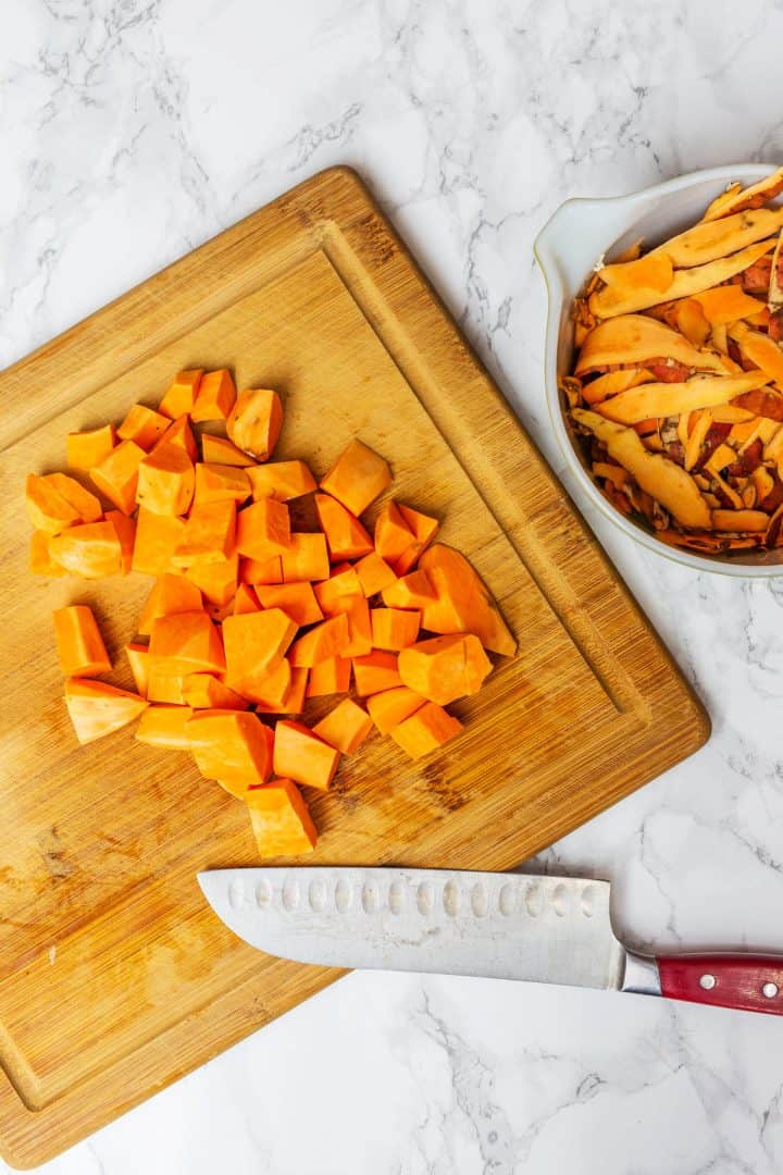 Chopped sweet potatoes on cutting board with knife and peels in bowl off to side