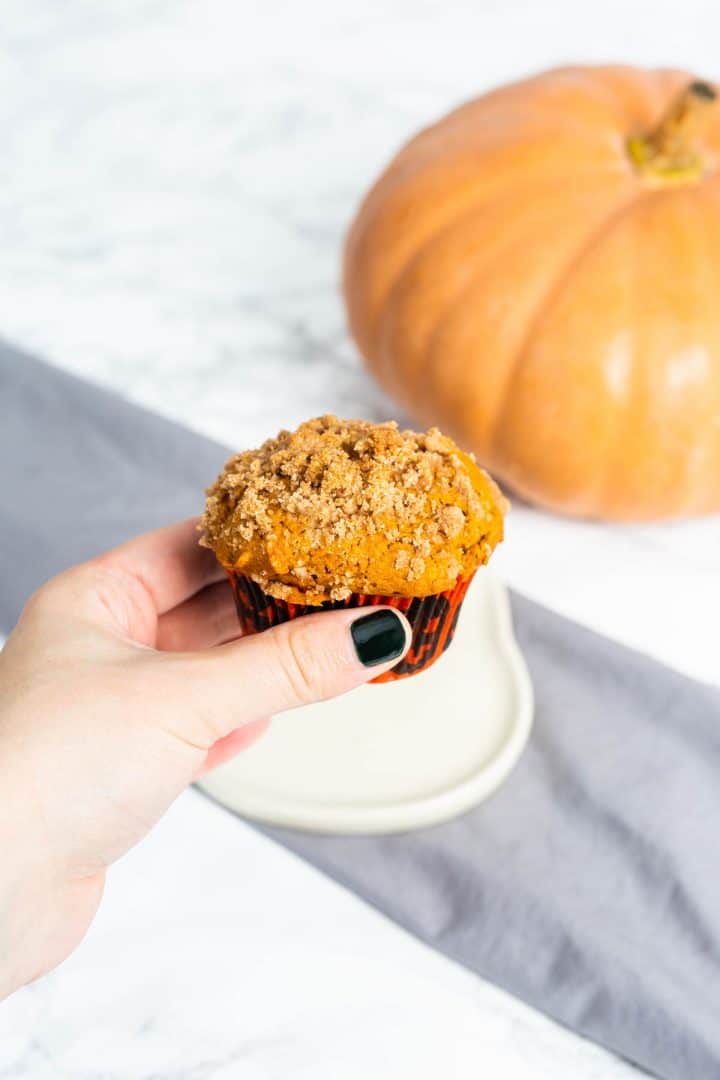 Hand holding pumpkin muffin over plate with cloth napkin and pumpkin in background
