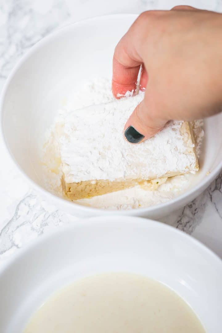 Hand dipping slab of tofu in bowl of cornstarch