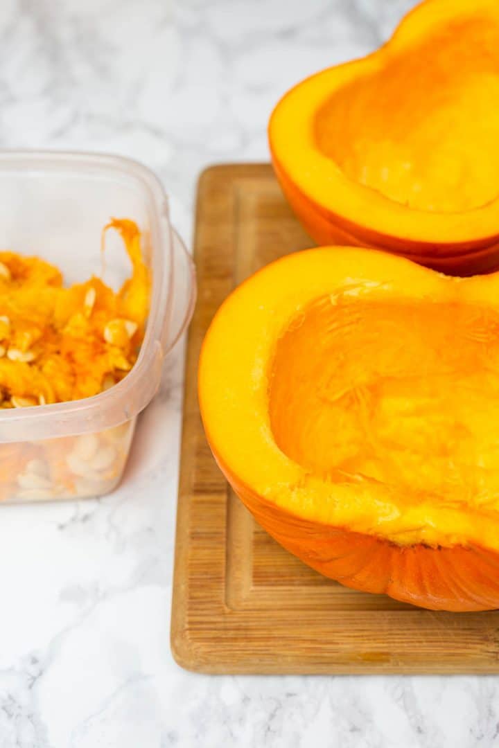Two pumpkin halves on cutting board with flesh and seeds in container off to side