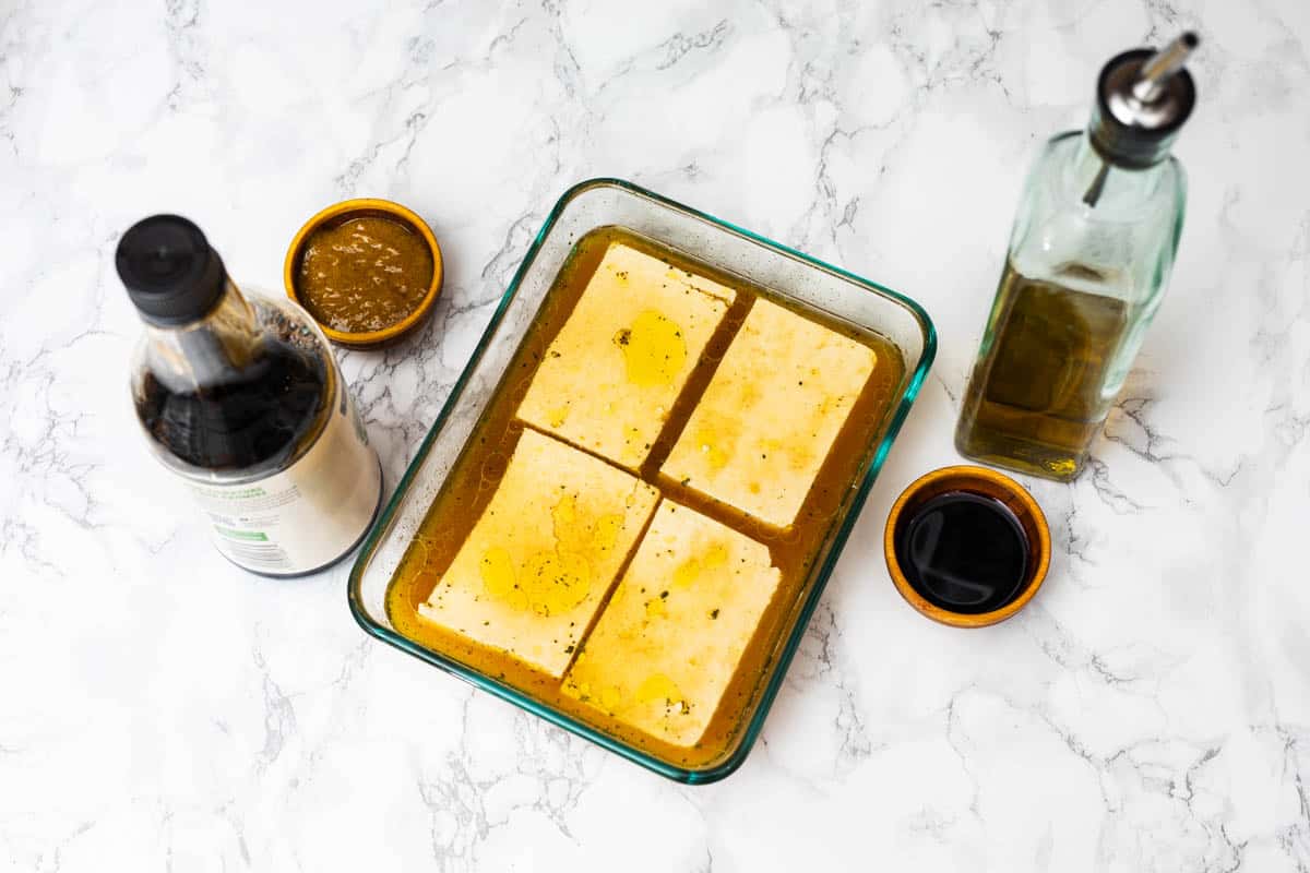Four pieces of tofu in marinade in glass dish with mustard, soy sauce, and olive oil off to side