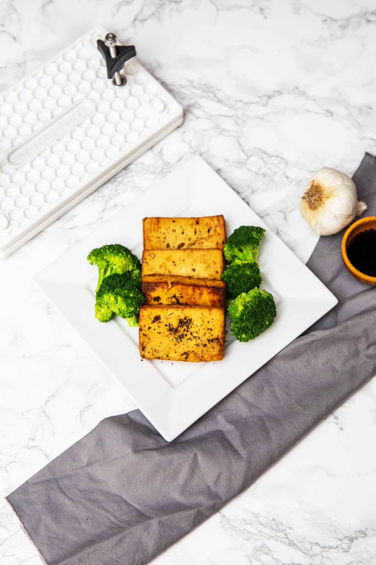 Four pieces of baked tofu on plate with broccoli with cloth napkin, garlic, soy sauce, and tofu press off to side