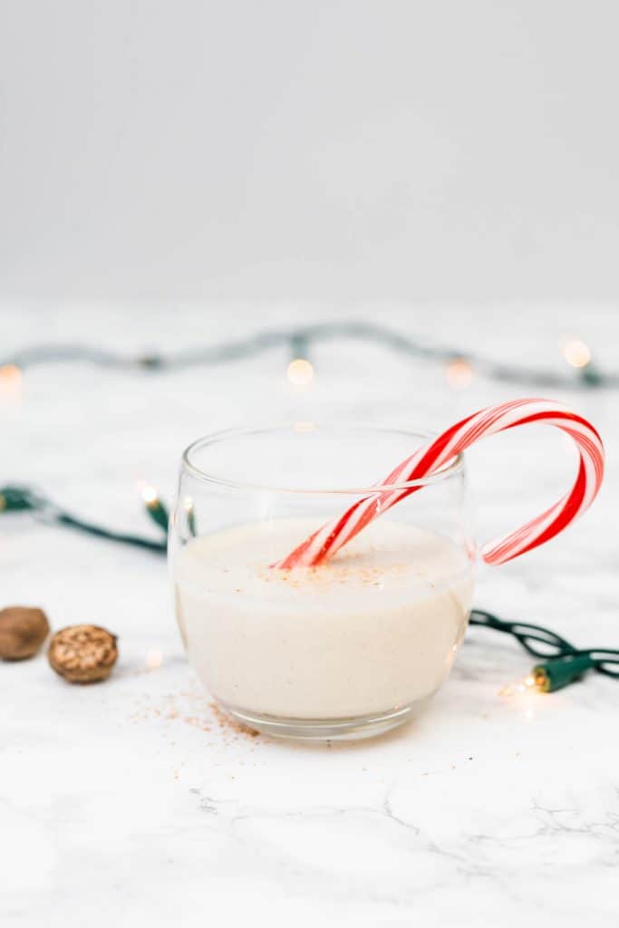 Glass of vegan eggnog with candy cane on marble counter with Christmas lights decor