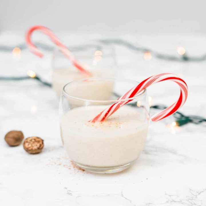 Two glasses of vegan eggnog with candy canes on marble counter with Christmas lights decor