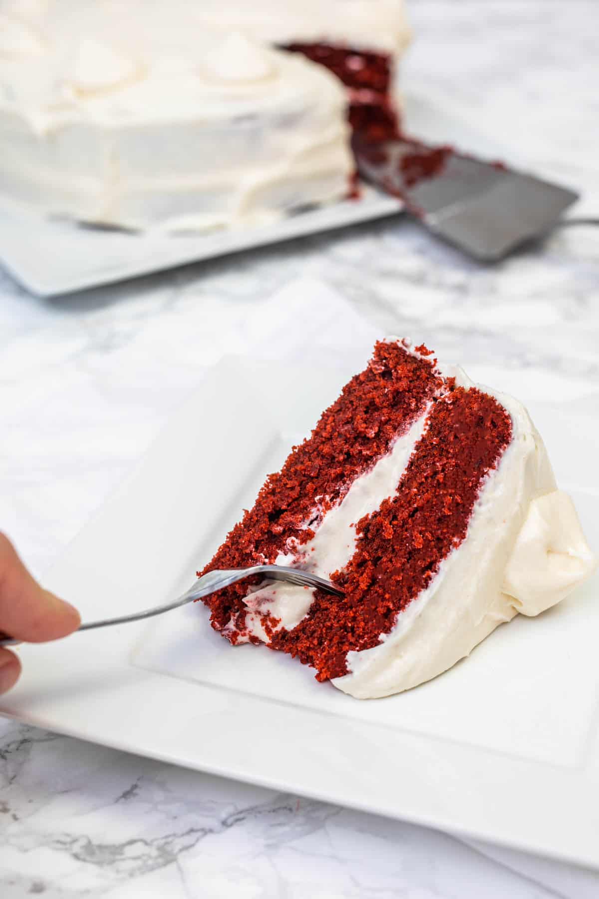 Hand sticking fork into slice of vegan red velvet cake with cream cheese frosting on white plate with cake in background