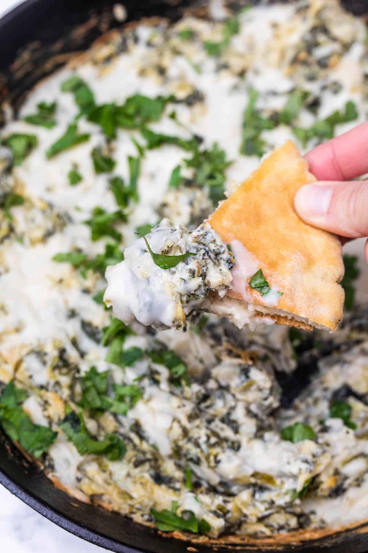 Hand scooping baked vegan spinach artichoke dip out of cast iron pan with piece of pita