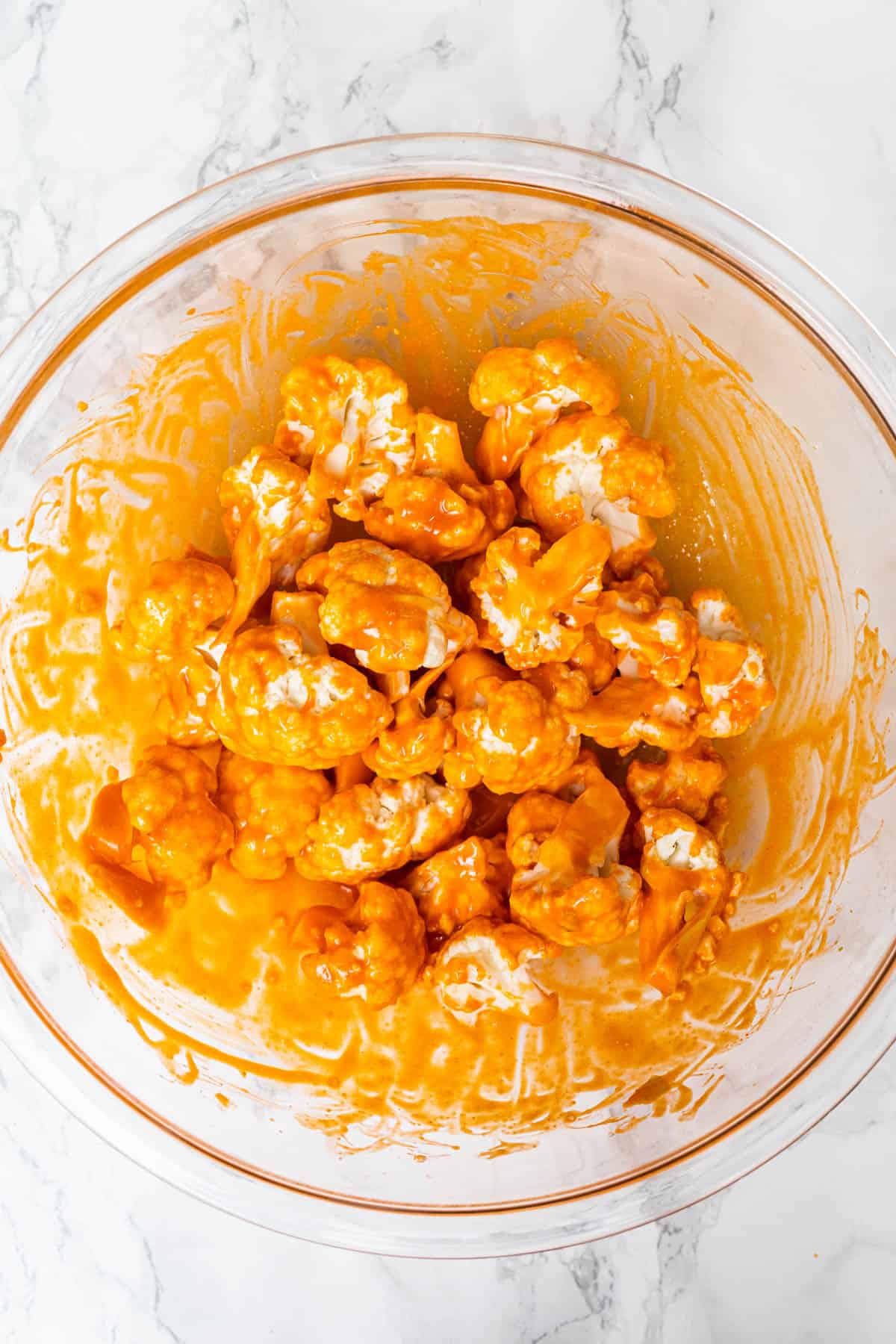 Large glass mixing bowl filled with raw cauliflower florets coated in Buffalo sauce