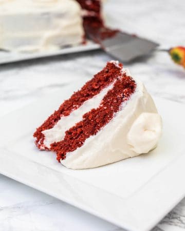 Slice of vegan red velvet cake with cream cheese frosting on white plate with cake in background