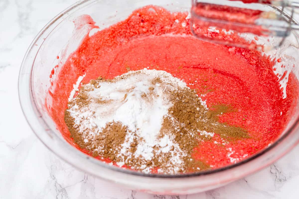 Red cake batter with cocoa powder and other ingredients on top in large glass mixing bowl on white marble counter