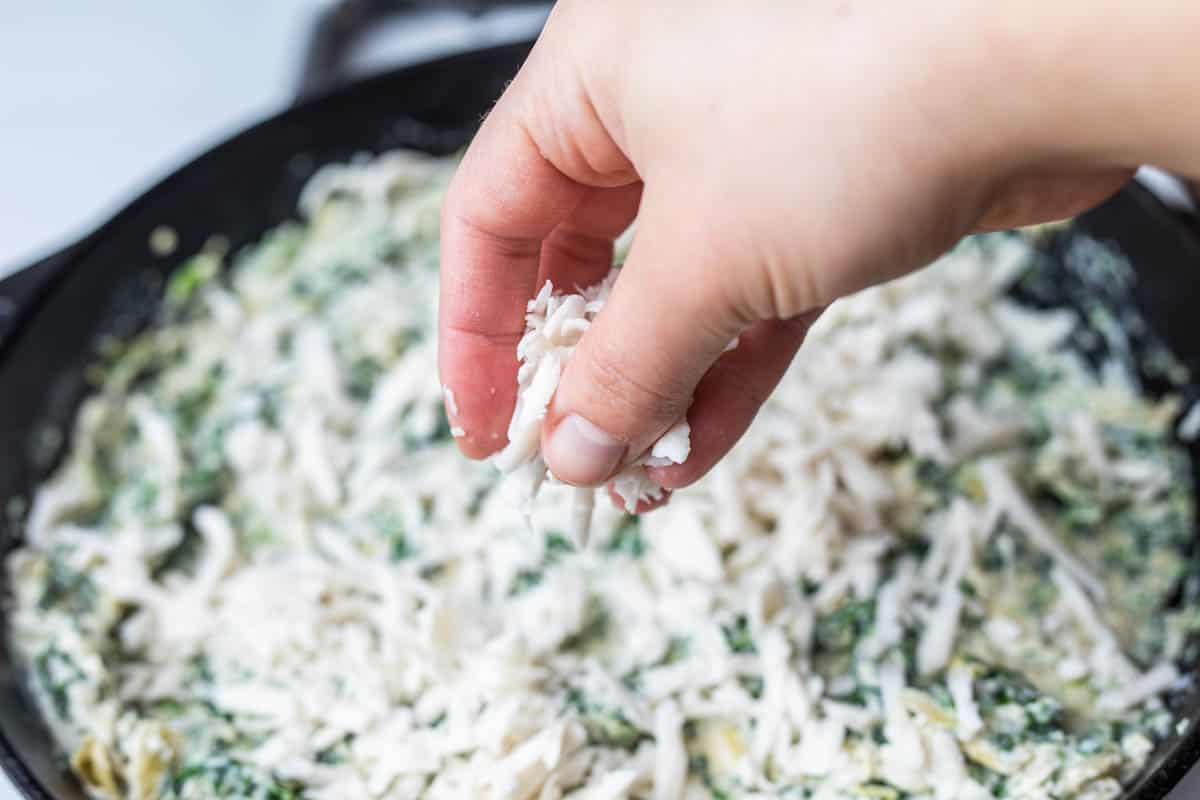 Hand sprinkling shredded vegan mozzarella cheese into unbaked spinach artichoke dip in cast iron pan