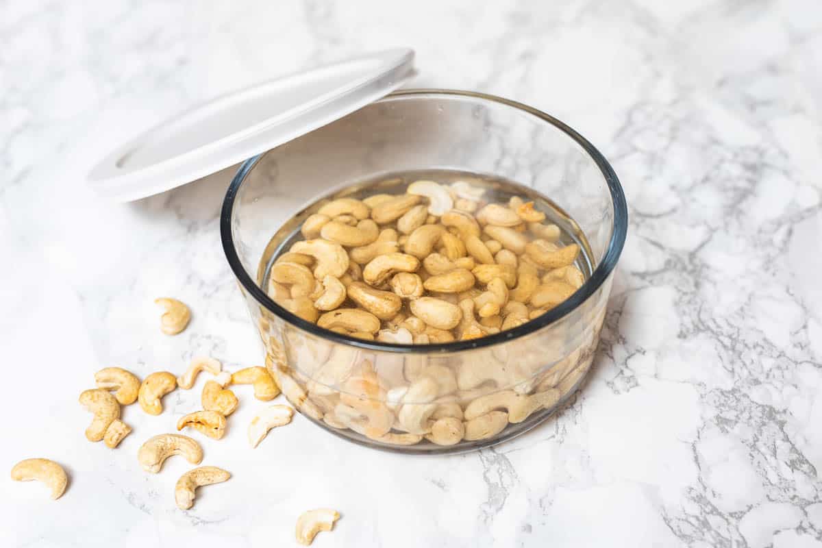 Raw whole cashews soaking in water in glass container with lid off to side