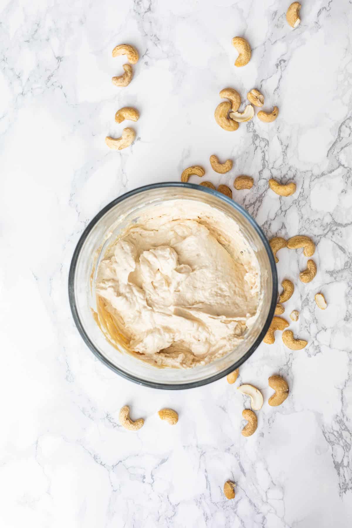 Cultured cashew cheese in glass dish surrounded by whole cashews on white marble surface
