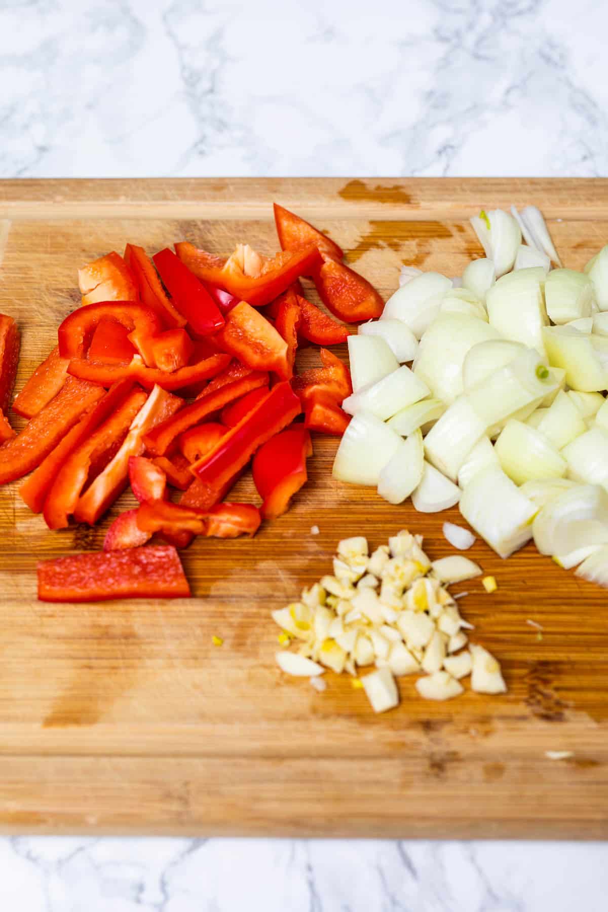 Sliced red bell peppers, onions, and garlic on bamboo cutting board on marble surface