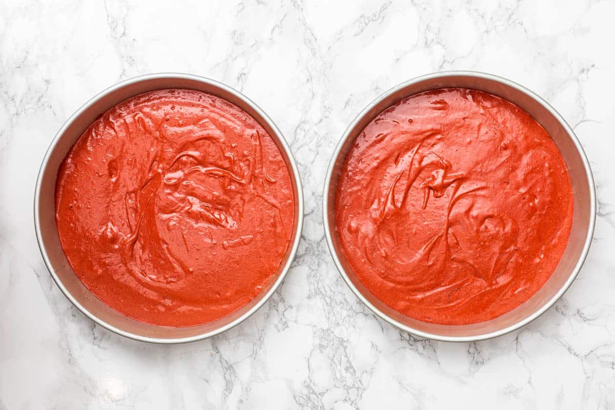 Two round cake pans filled with vegan red velvet cake batter on white marble surface