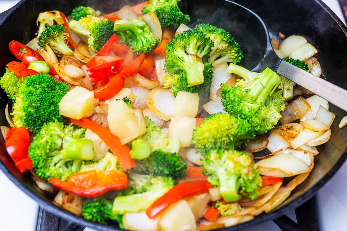 Broccoli, onions, red bell peppers, and pineapple cubes being stir fried in cast iron pan