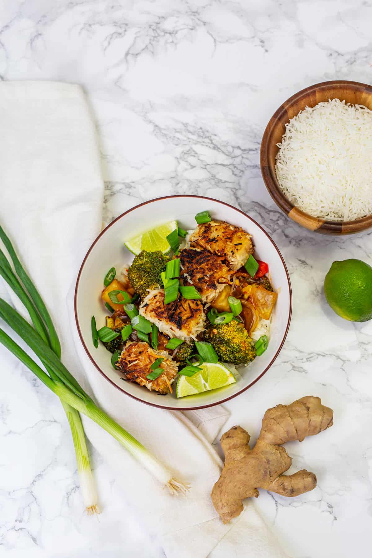 Bowl of coconut crusted tofu stir fry with lime, bowl of shredded coconut, green onions, and ginger root surrounding bowl