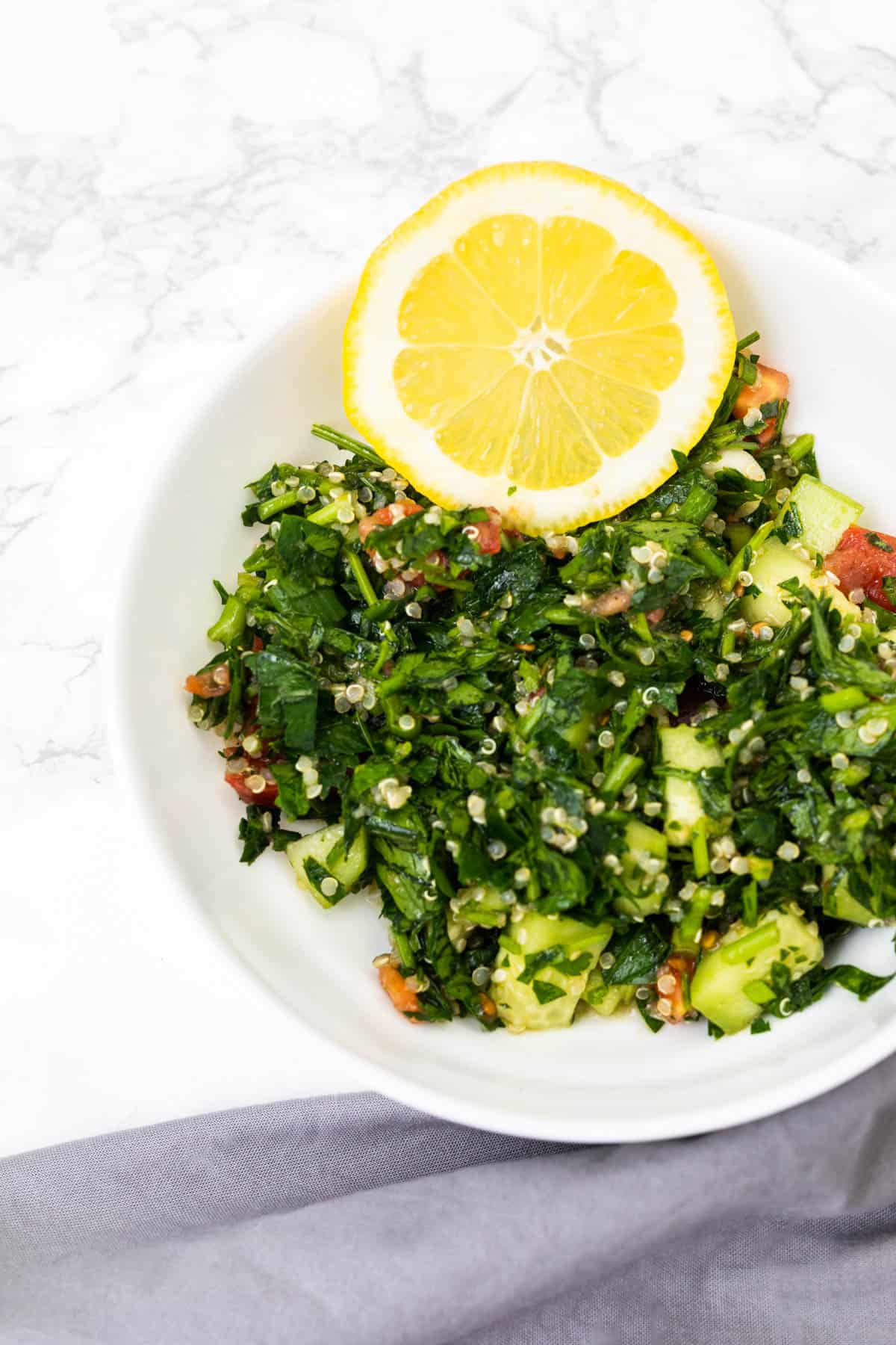 Bowl of quinoa tabouli with lemon slice and gray cloth napkin on white marble counter
